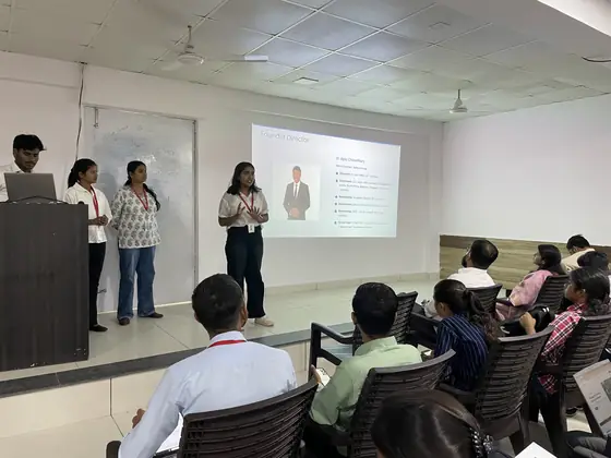 A team presenting to a seated classroom beside a slide introducing SSS's founder director